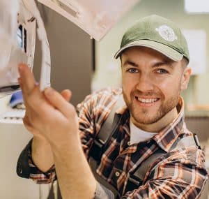 Handyman with a checkered shirt with a white under shirt and a green hat. Working on a AC unit