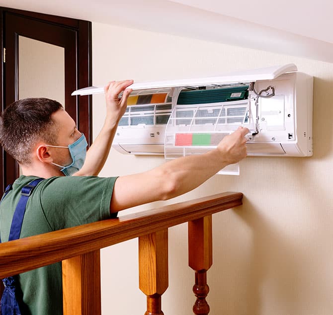 Handyman waring green and blue and a mask working on a AC unit inside