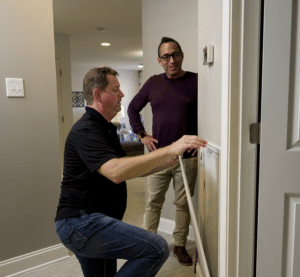 Two men talk while one kneels to adjust a thermostat on a wall in a modern home interior.