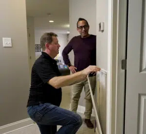 Two men talk while one kneels to adjust a thermostat on a wall in a modern home interior.