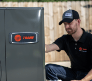 A technician kneels beside a Trane HVAC unit outdoors, inspecting or servicing the equipment.