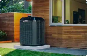 A large outdoor HVAC unit sits on a platform next to a wooden house with a window.