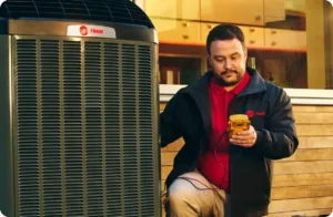 Technician in a red shirt checks a large outdoor Trane AC unit with a yellow meter.