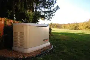 A Generac standby generator sits on gravel near a brick wall beside a grassy yard with trees in the background.
