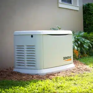 A Generac standby generator installed outside a house on a mulched area near green grass and plants.